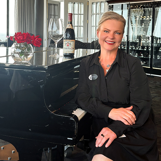 Woman sitting at a piano with wine and glassware, wearing a black outfit, smiling in an elegant restaurant setting.