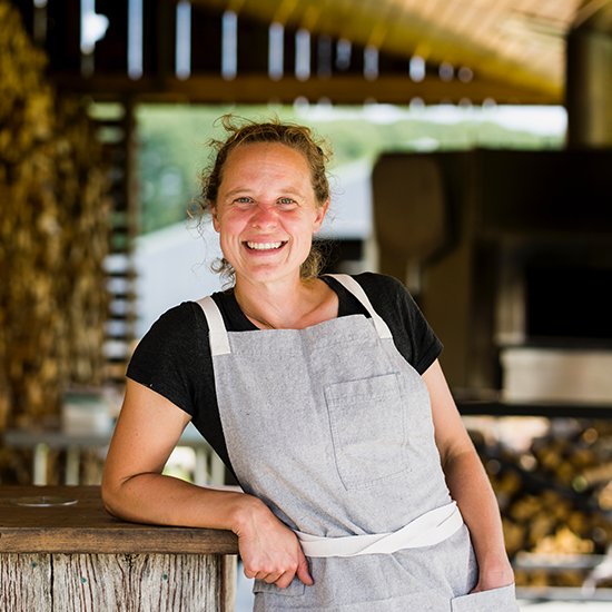 Woman standing outdoors in front of a wood-fired oven, wearing a gray apron and smiling.