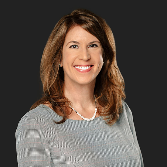 Professional headshot of a smiling woman with shoulder-length brown hair wearing a gray patterned blouse and necklace on a dark background.