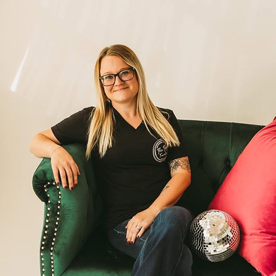 Woman seated on a green couch with bright pink pillows and a disco ball, wearing glasses, jeans, and a black shirt.