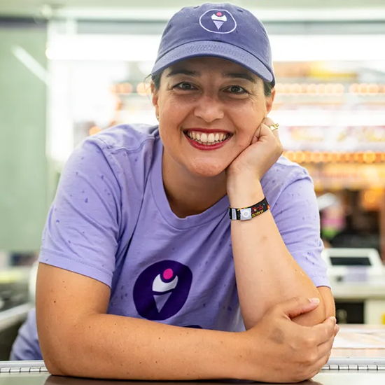 Smiling woman wearing a purple hat and t-shirt with an ice cream logo, leaning on a counter in a casual setting.