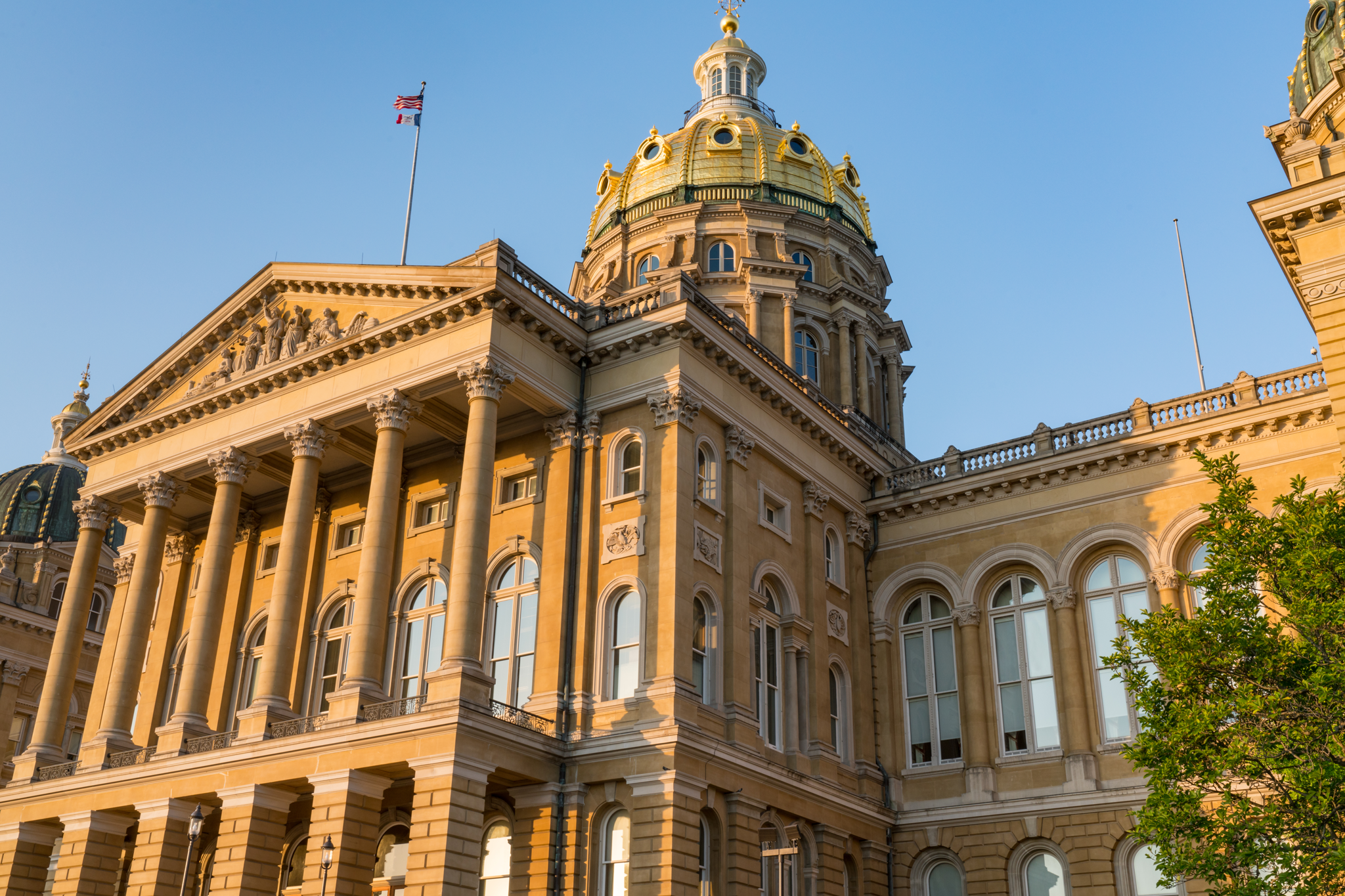 Exterior view of the Iowa State Capitol building with golden dome and American flag against a clear blue sky.