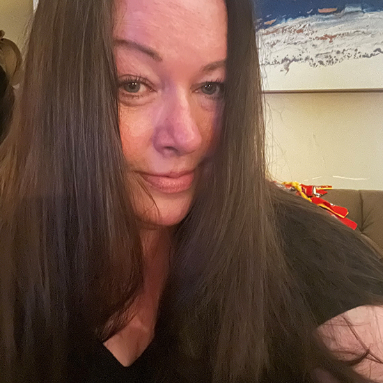 Close-up photo of woman with long brown hair smiling while wearing a black top indoors.