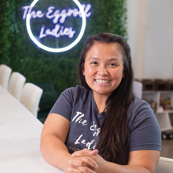 Smiling woman seated at a table wearing a ‘The Eggroll Ladies’ t-shirt with a neon sign reading ‘The Eggroll Ladies’ in the background.