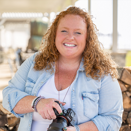 Smiling woman with curly red hair in a denim shirt holding a camera outdoors.